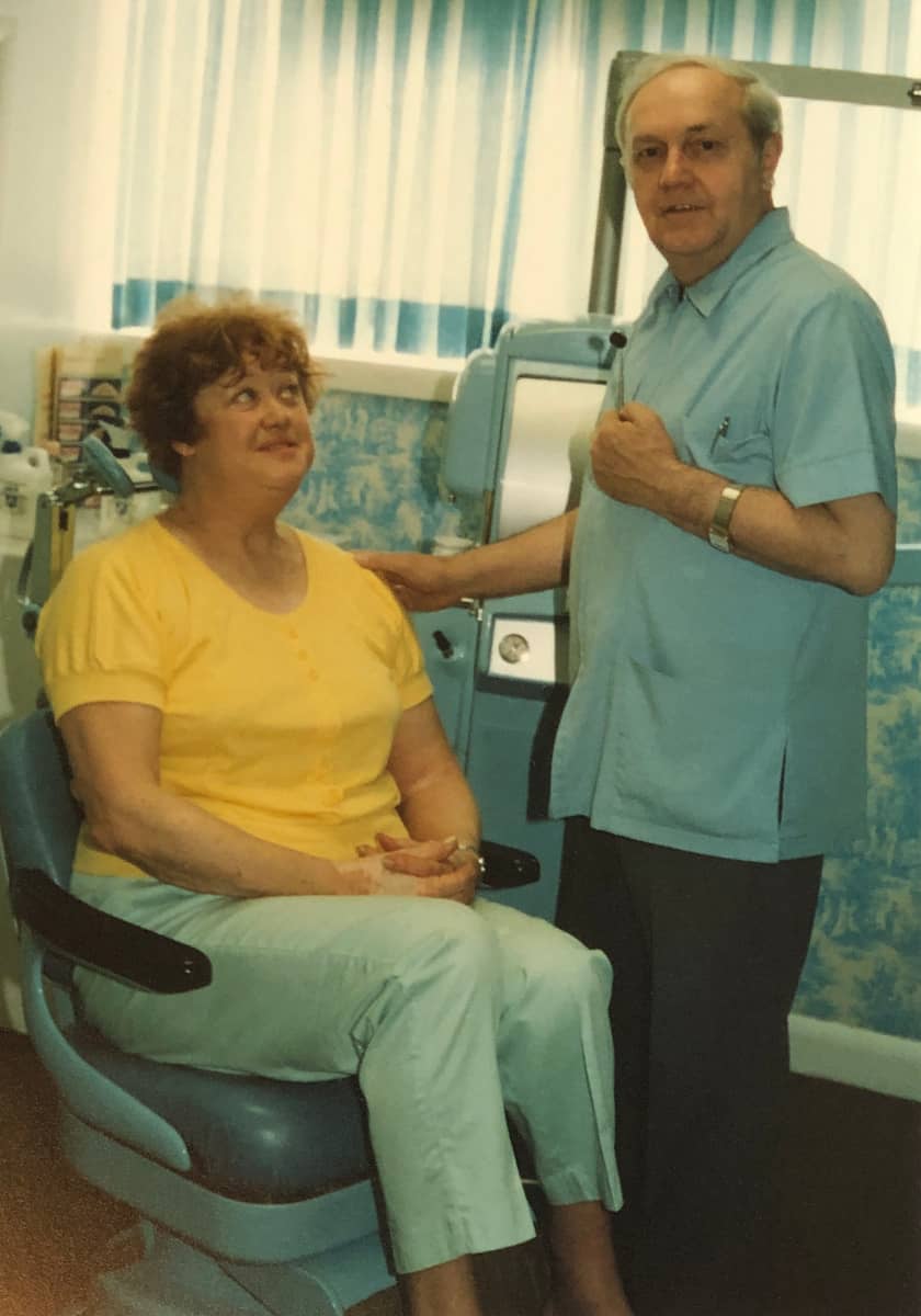 Denturist Joe Schankula posing facing the camera with his arm gently touching the shoulder of a female patient in the dental chair looking up at him