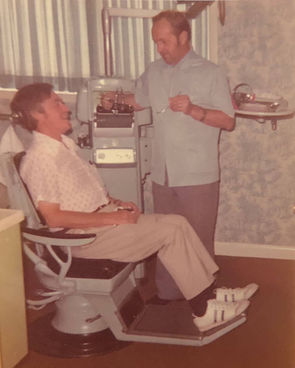 Denturist Joe Schankula holding an oral mirror, with a smiling male patient in the dental chair