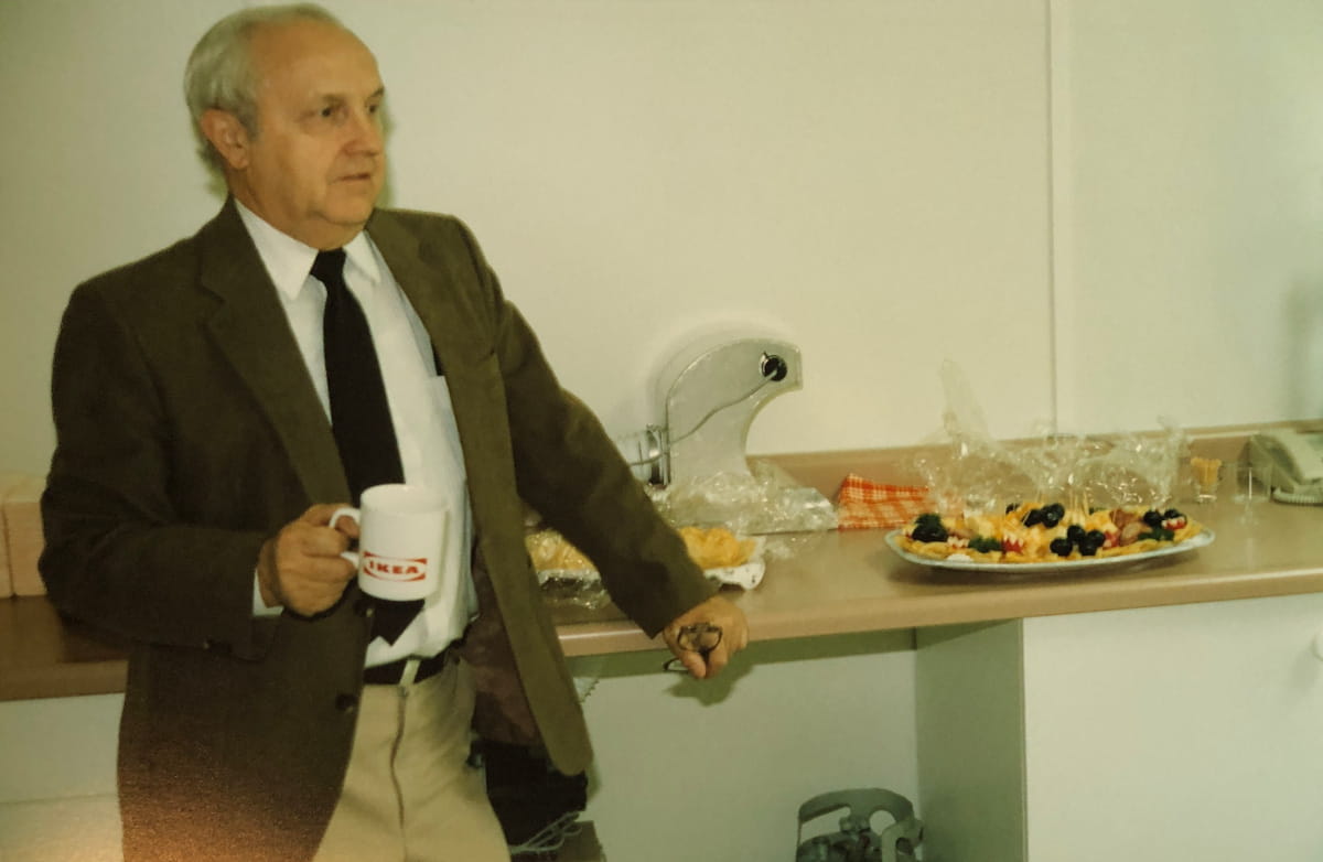 Denturist Joe Schankula at his son Tim's grand opening of his Oakville Denture Clinic, in a suit, holding a mug, with hors d'oeuvres beside him on the lab countertop
