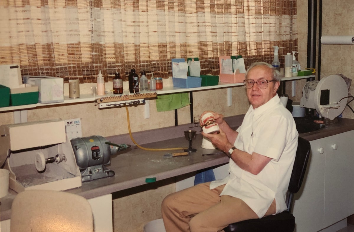 Denturist Joe Schankula, Tim's father, in the lab holding a set of dentures on the articulator, with a calendar showing May 1993
