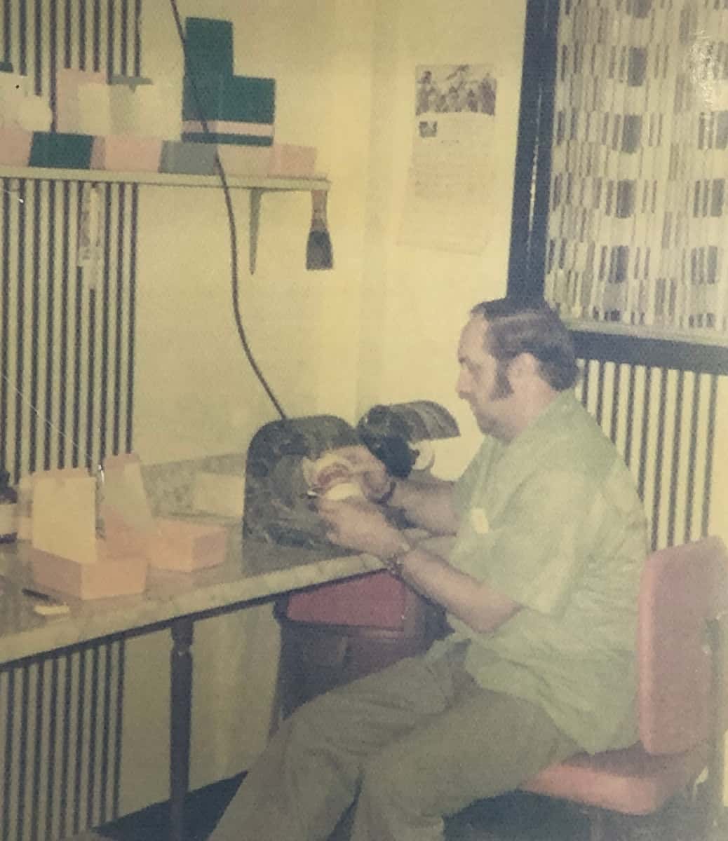 Denturist Joe Schankula in the lab at the downstairs Grantham Denture Clinic looking at a set of dentures in an articulator in his hands, circa 1970s