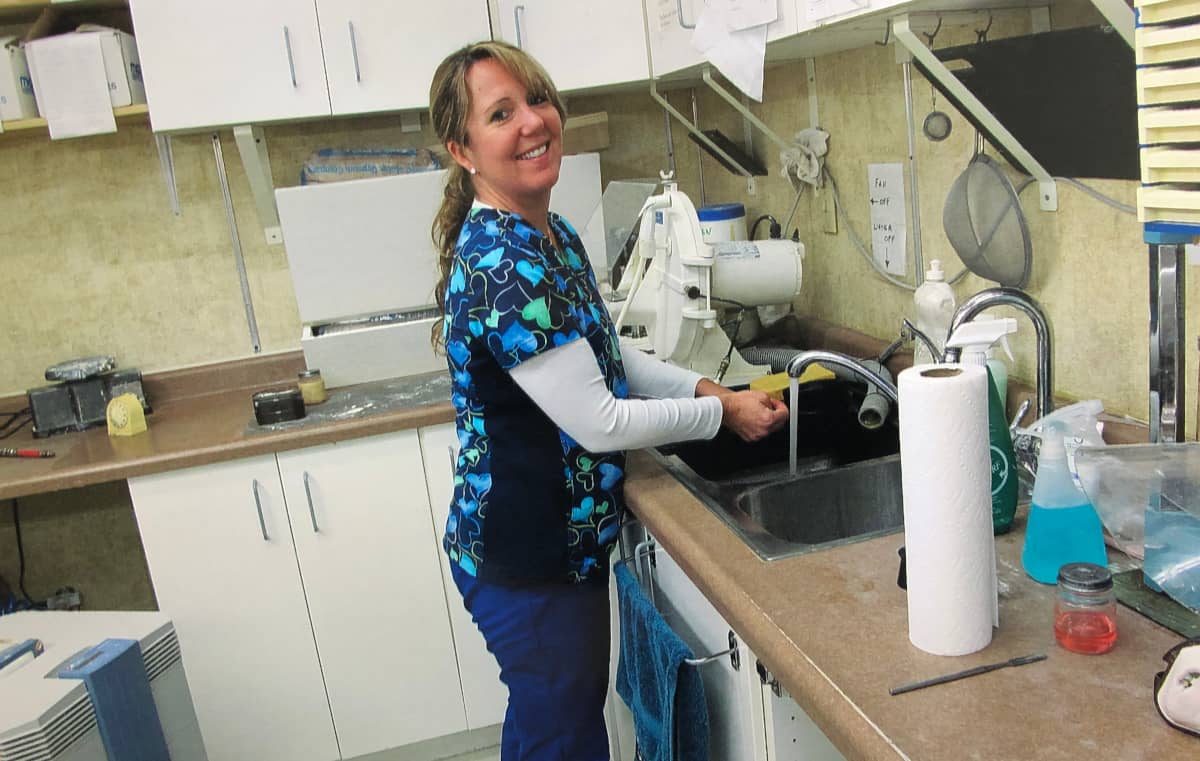 Receptionist and lab assistant, Linda, at the sink in the lab with the tap running, smiling at the camera