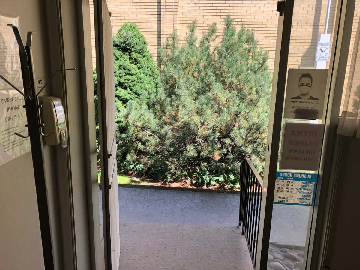 A patient's point of view about to step through the opened door to the porch before them, a railing on their right and the big garden bushes in front of them