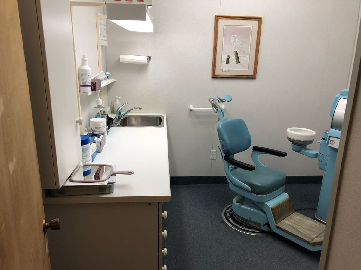 The operatory with its sterilized counter and sink on one side and the dental chair and spit bowl on the other, showing wildlife artwork of an owl in winter and papertowels mounted on the wall, and containers of impression material and a handheld mirror on the countertop
