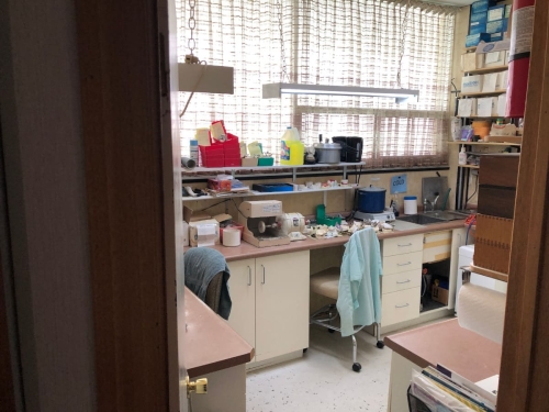 Shot into the on-site denture lab from the lab's doorway, showing wall-to-wall curtained windows, cabinets, drawers and shelves with dentures, equipment and supplies, two chairs on rollers, one with a dental gown draped over it, a fire extinguisher mounted on the wall inside the doorway and lights hanging from chains from the ceiling to augment the ceiling lights