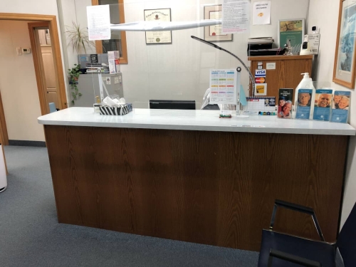 Closeup of the reception desk from inside the entrance, showing its mandated plexiglass protective barrier, a box of tissues, flip calendar and pamphlets on the countertop, a full-year calendar on a sheet of paper taped to the plexiglass, diplomas on the wall behind reception and a metal filing cabinet on one side and a wooden cabinet on the other