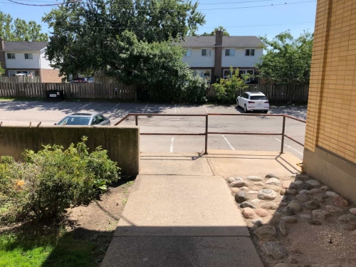 The end of the garden alleyway at the rear of the plaza, showing some bushes, sidewalk and staircase railing going down to the rear parking lot showing a couple of parked cars and backyards of houses