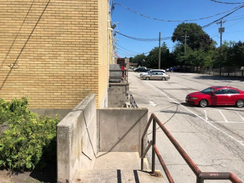 The parking lot at the rear of the Grantham Plaza as seen from the top of a stone staircase at the end of the Denture Clinic's garden alleyway, looking towards the Niagara Street entrance, with a number of steps down, steel railing, some garden alleyway bushes on the left and a few parked cars below