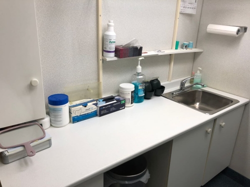Clean and tidy countertop and sink, with wall-mounted papertowels, a little shelf with tissues and dixie cups, cupboards, and with a handheld mirror, containers with impression material, boxes of dental gloves, a large pump of Listerine mouthwash and impression mixing rubber bowls on the countertop