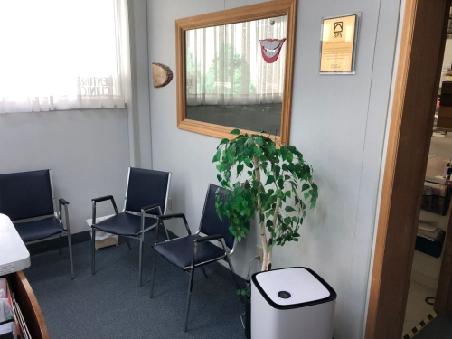 31-A shot from the reception desk of the waiting room, with a mirror on the wall, an artificial plant and air purifier on the floor in the foreground, and a tiny sliver showing into the lab through the doorway on the right edge