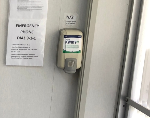 Closeup of the hand-sanitizer dispenser mounted on the wall just inside the door, next to a sheet taped on the wall showing "Emergency Phone Dial 9-1-1" and location information and directions to give to first responders