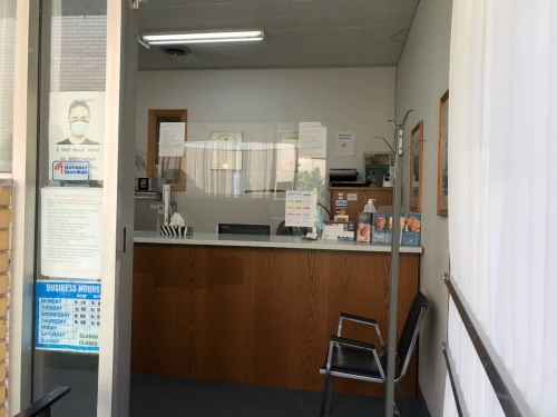 A patient's point of view crossing our threshold into our reception area, showing more of the reception desk with its mandated plexiglass protective barrier, and a coat tree, a chair, and pamphlets on our reception desk countertop